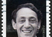 U.S. Navy Ship to Be Named After Harvey Milk A photo of a postage stamp with Harvey Milk's portrait on it. The photo is in black and white, aside from a small rainbow in the upper left-hand corner.