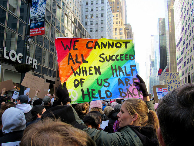 A photo of a female protestor holding a sign that reads, "We cannot all succeed when half of us are held back."