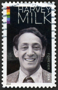 A photo of a postage stamp with Harvey Milk's portrait on it. The photo is in black and white, aside from a small rainbow in the upper left-hand corner.